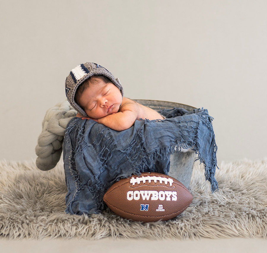 A newborn is captured in a bucket with a football ball by a photographer in Dallas-Fort Worth, Texas.